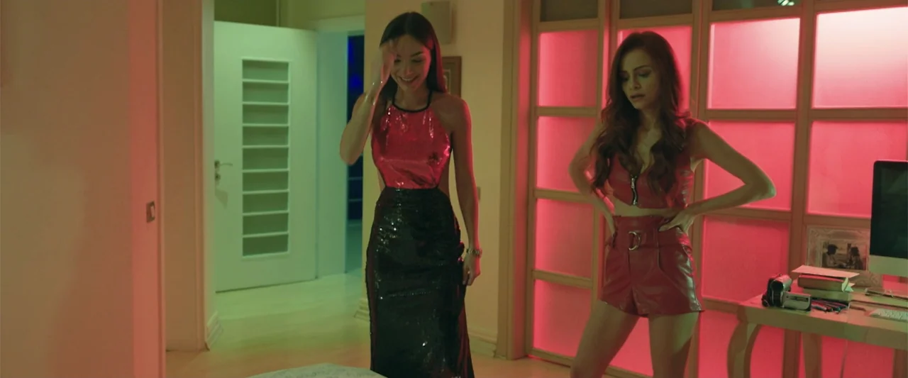 Wrong Package (2025) movie still: fullbody — A modern room features a grid-patterned glass screen glowing with bright pink light. Two women stand in…