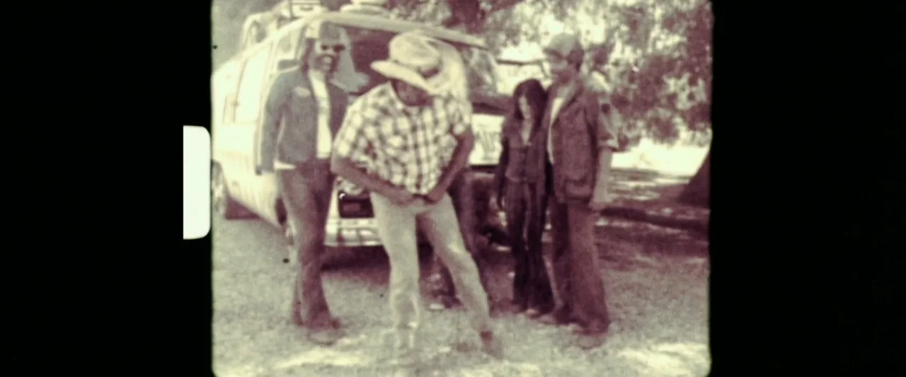 31 (2016) movie still: fullbody — A group of people stands outside a van parked in a shaded outdoor area. One man in a cowboy hat adjusts his belt…