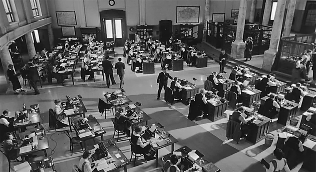 Kafka (1991) movie still: establishing — A vast, high-ceilinged office space features rows of wooden desks and industrial columns. Numerous clerks…
