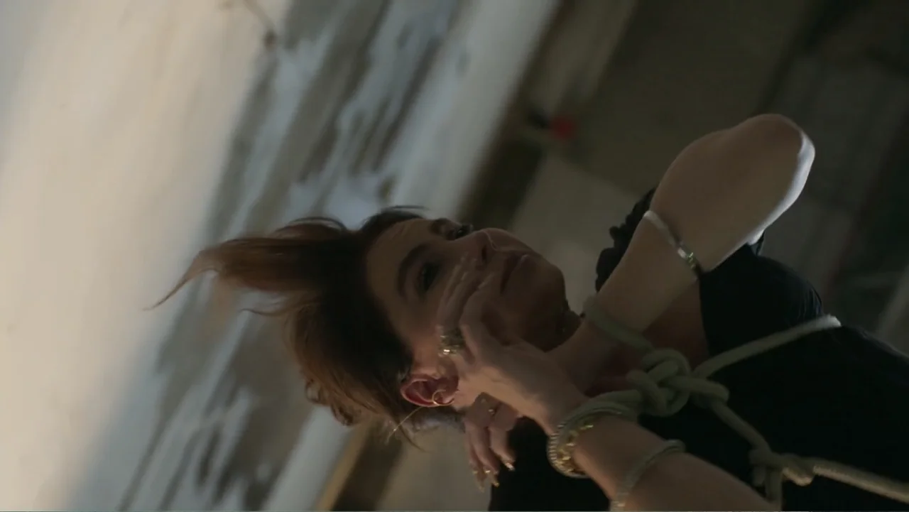 Making of: Scars of Beauty (2025) movie still: medium — A dark industrial interior with blurred concrete walls visible in the background. A woman in…