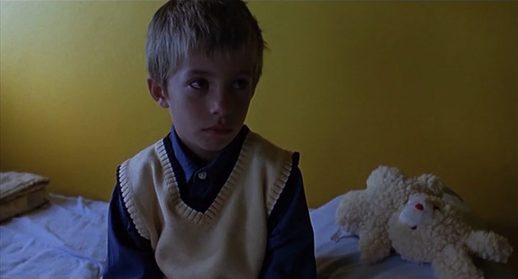 The Heart Is Deceitful Above All Things (2004) movie still: medium — A young boy sits on a bed against a solid yellow wall. A plush white stuffed…