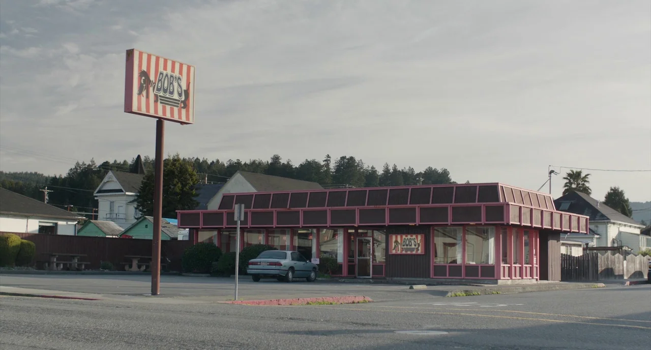 An Evening With Beverly Luff Linn (2018) movie still: establishing — A roadside diner with pink and brown trim sits in a mostly empty lot. A tall,…