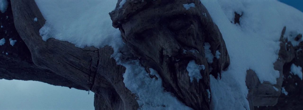 The Hateful Eight (2015) movie still: closeup — A weather-worn wooden crucifix stands outdoors during a cold winter twilight. The carved face of the…