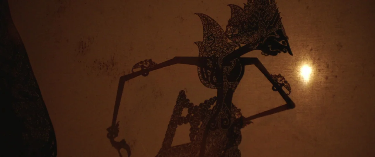 Impetigore (2019) movie still: closeup — A detailed leather wayang kulit puppet silhouette is projected onto a translucent fabric screen. The figure…