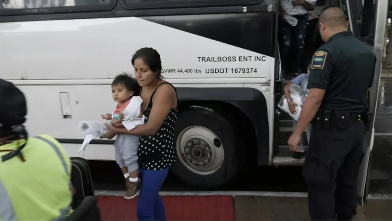 Torn Apart: Separated at the Border (2019) movie still: medium — An outdoor bus boarding area features a large white passenger bus parked on…