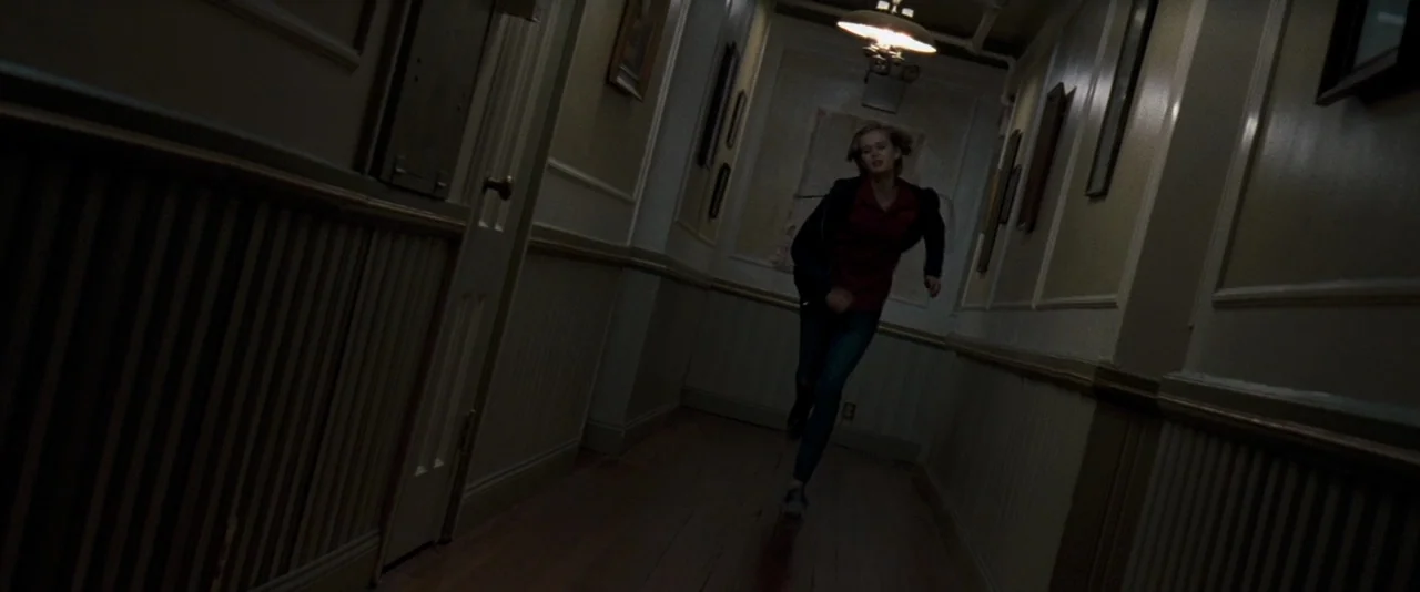 The Innkeepers (2011) movie still: fullbody — A dim residential hallway features paneled wainscoting and several small framed pictures on walls. A…