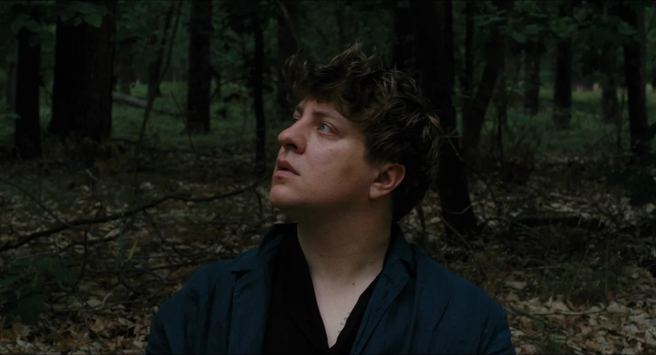 Afire (2023) movie still: medium — A dense, dark woodland area with scattered leaves and thin tree trunks. A young man stands in the clearing…