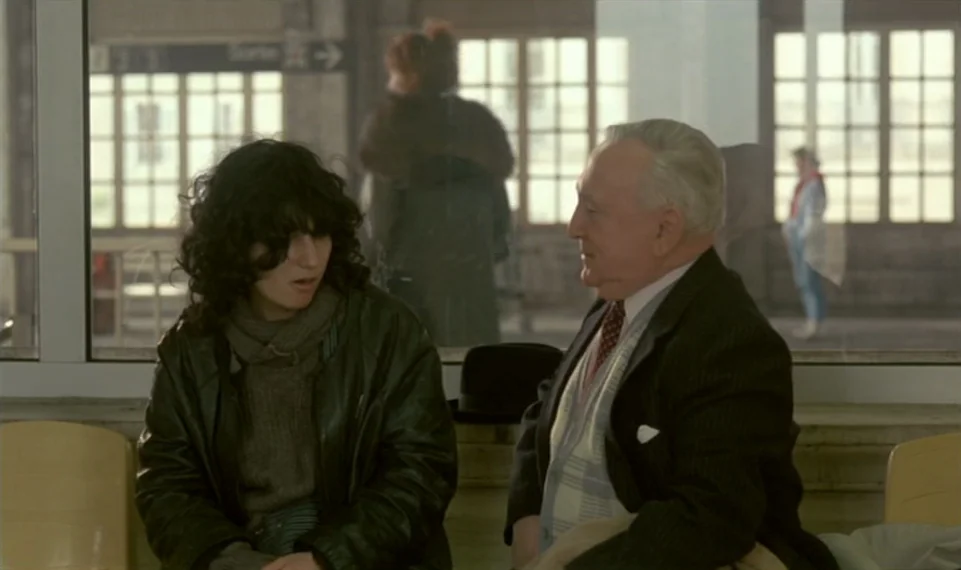 Vagabond (1985) movie still: medium — An airport or train terminal waiting area with large grid-patterned windows behind them. The two people sit on…