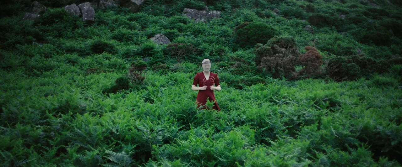 Fréwaka (2024) movie still: wide — A dense hillside is covered in vibrant green ferns and scattered gray rocks. A lone person wearing a red dress…