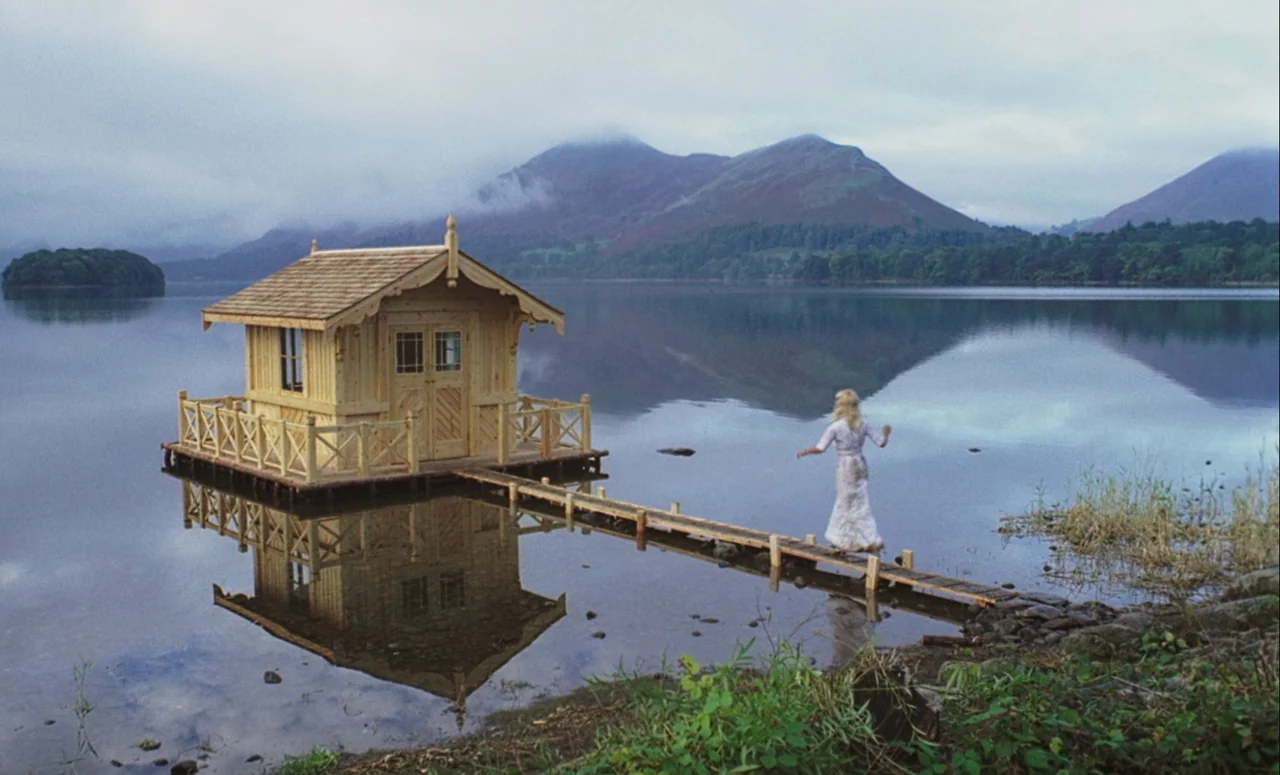 Mahler (1974) movie still: wide — A calm lake features a small, light-wood structure floating atop the water. A woman in a white dress walks across…