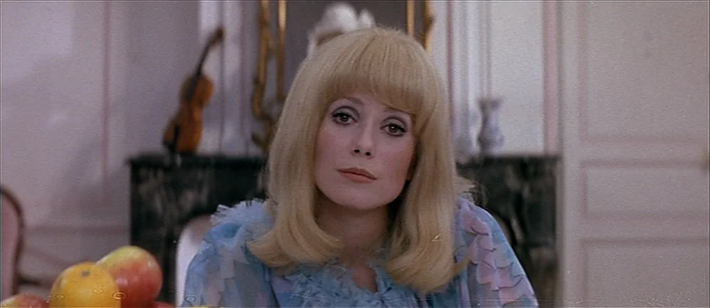 Les Demoiselles de Rochefort (1967) movie still: closeup — A light-toned indoor space features a dark marble fireplace with a violin on top. The…