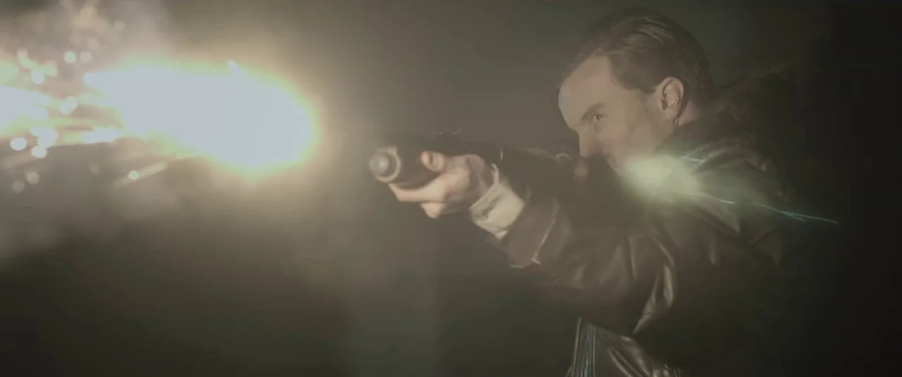 The Void (2016) movie still: medium — A man in a leather jacket stands in darkness holding a firing rifle. A large, bright muzzle flash illuminates…