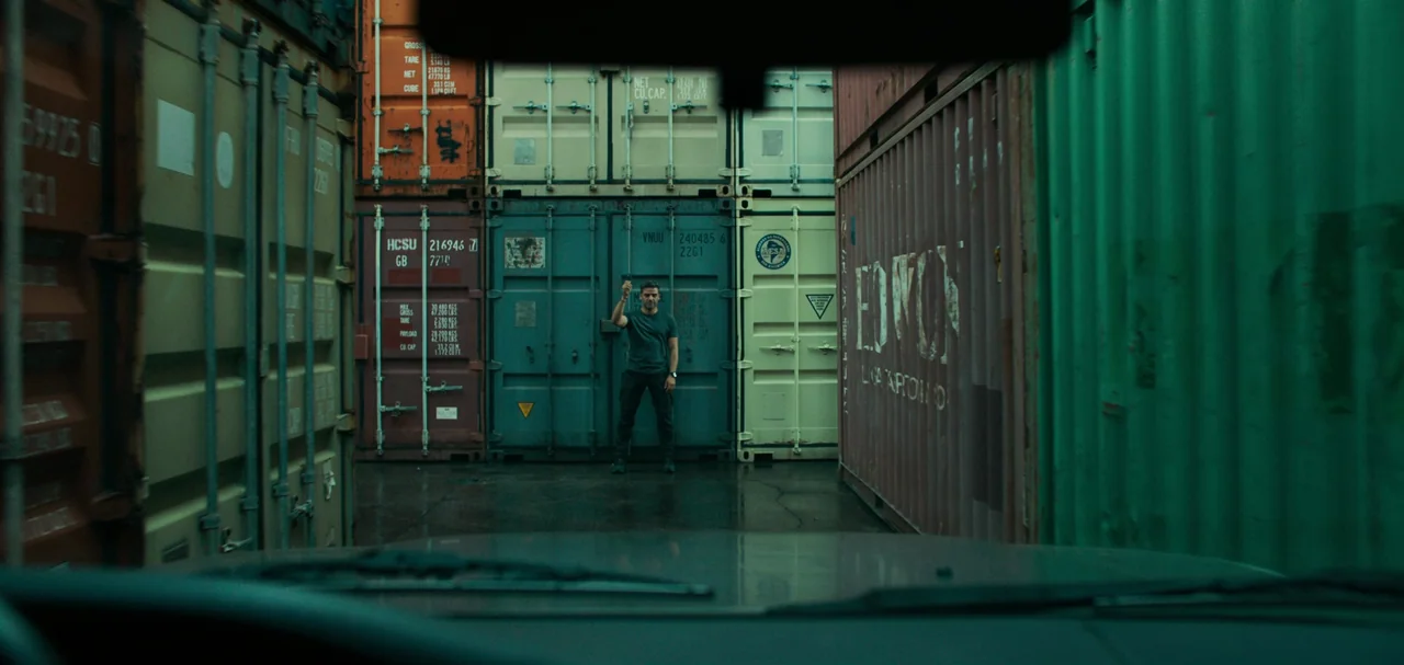 Triple Frontier (2019) movie still: wide — A man stands between stacks of colorful shipping containers in an industrial yard. The view is framed…