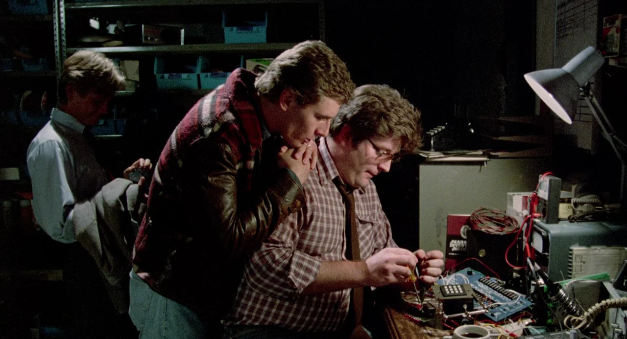 Crimes of Passion (1984) movie still: medium — A dimly lit room filled with technical equipment and cluttered shelves of spare parts. Two men work…