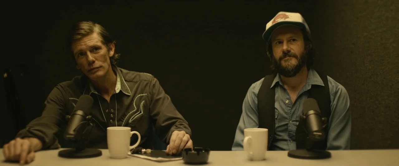 Blaze (2018) movie still: medium — Two men are seated at a table against a solid, dark soundproof wall. They are positioned behind professional…
