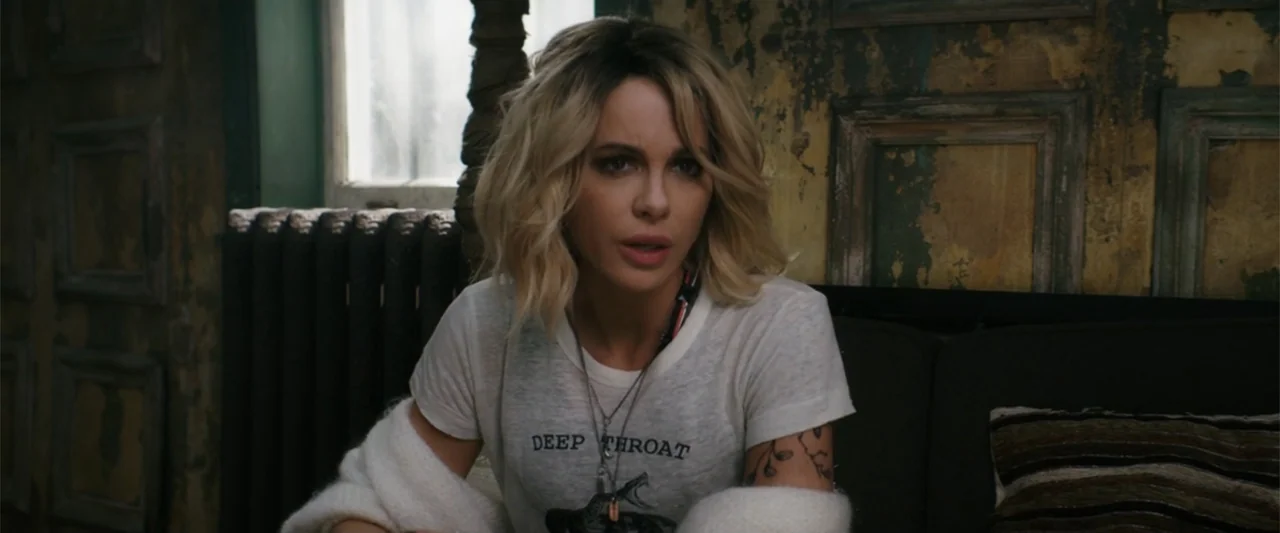 Jolt (2021) movie still: medium — A room with distressed wall panels and a black radiator near a window. A woman in a graphic t-shirt looks…
