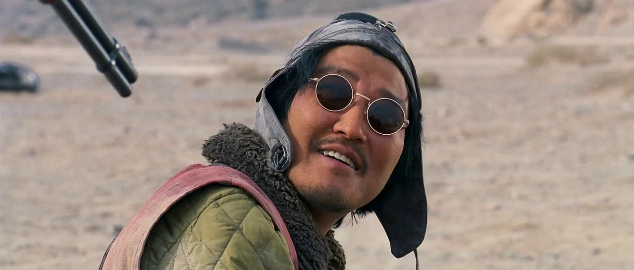 The Good The Bad The Weird (2008) movie still: closeup — An open desert environment with sandy ground and blurred terrain in the background. A man…