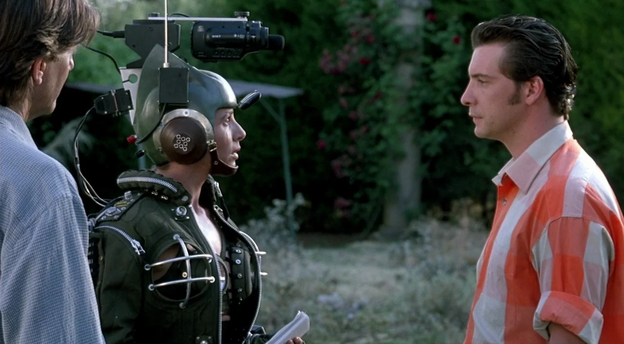 Kika (1993) movie still: medium — An outdoor area features dark foliage and a glimpse of a structure behind. A woman wearing a mounted helmet camera…