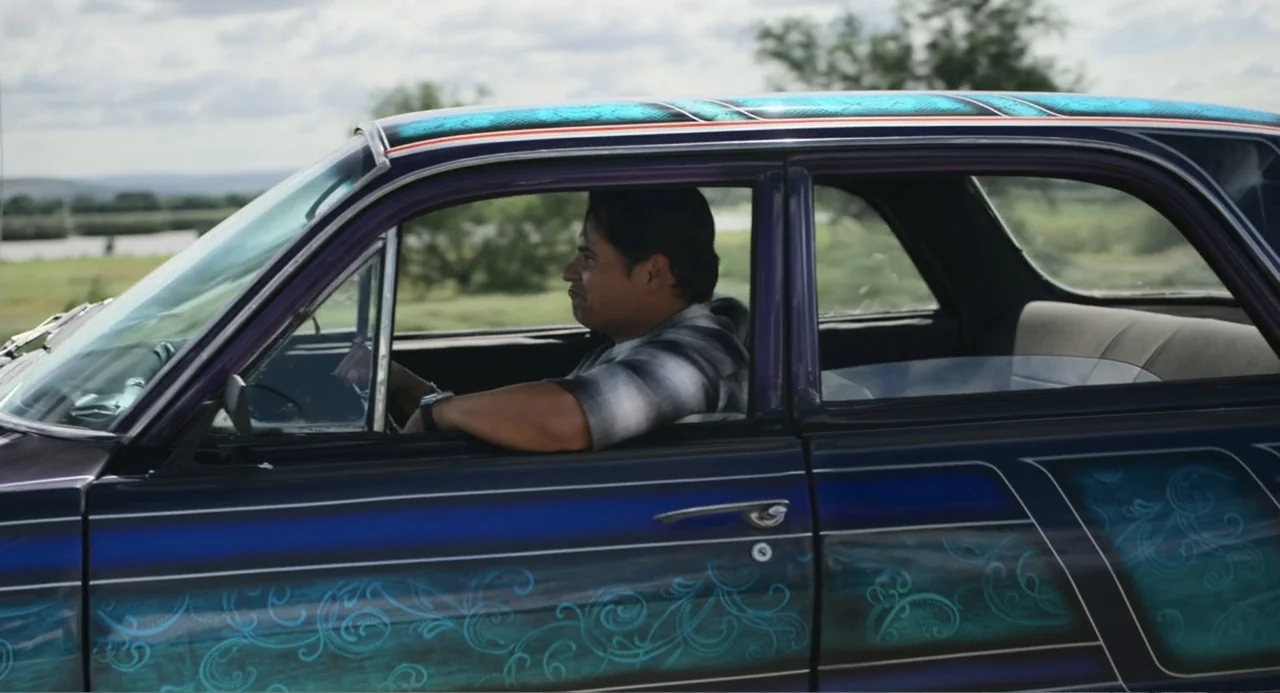 A Million Miles Away (2023) movie still: medium — A man drives a dark purple custom car with elaborate teal painted pinstriping patterns. The…