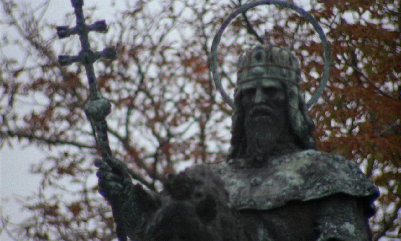 The Church (1989) movie still: medium — A bronze sculpture of a king with a halo stands in a public park. The figure holds a double-barred cross…