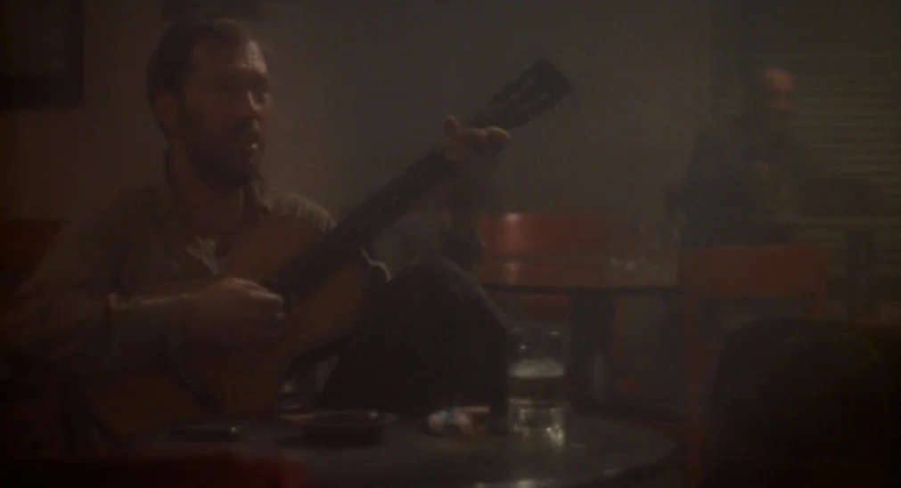 Bound For Glory (1976) movie still: medium — A dimly lit, smoke-filled space with round tables and chairs visible in darkness. A bearded man sits at…