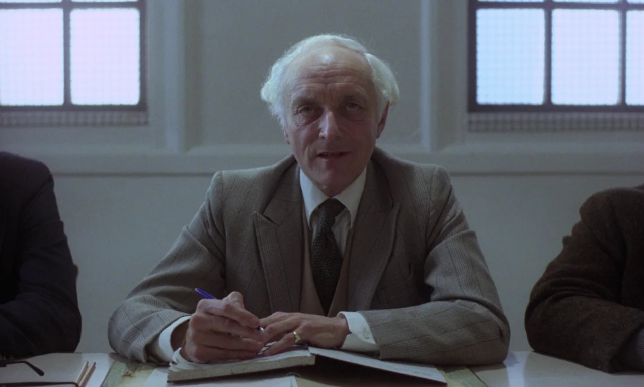 Scum (1979) movie still: medium — A spare, light-filled meeting room features plain walls and windows with visible gridded panes. A man sits at a…