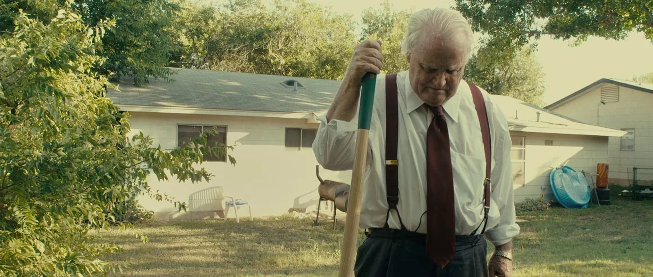 The Imposter (2012) movie still: medium — A residential backyard behind a single-story house features a patch of dry grass. An elderly man in formal…