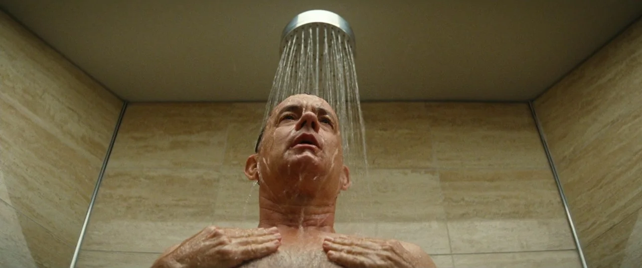 A Hologram for the King movie still: medium — A tiled enclosure features beige stone-patterned walls and a metal shower head above. A man stands…