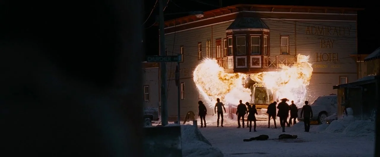 30 Days of Night (2007) movie still: wide — A snowy street in a dark town features an explosion engulfing a wooden hotel building. A group of…