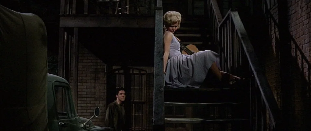 Wild in the Country (1961) movie still: wide — An outdoor brick building with a dark staircase at night. A woman sits on the stairs with a guitar…