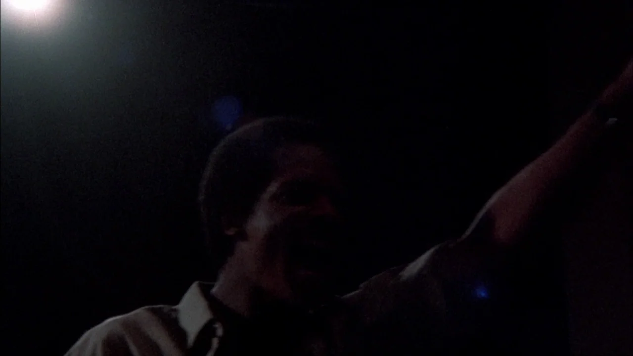 Stop Making Sense (1984) movie still: medium — A dark environment with a single bright light source casting shadows on a man. The man is raising one…