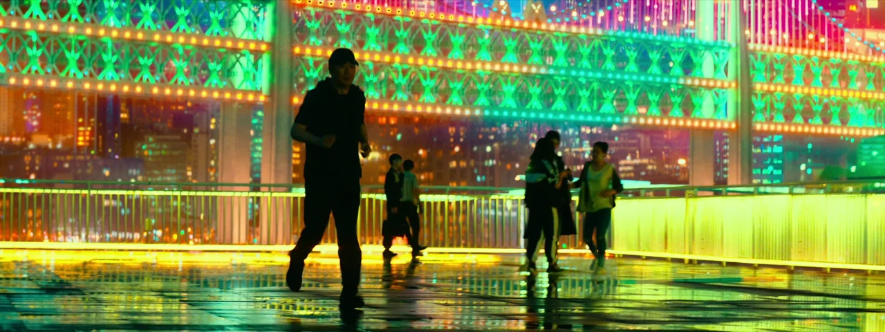 Under The Light (2023) movie still: wide — A wet, paved pedestrian walkway features bright, multi-colored neon bridge lighting overhead. A man in…