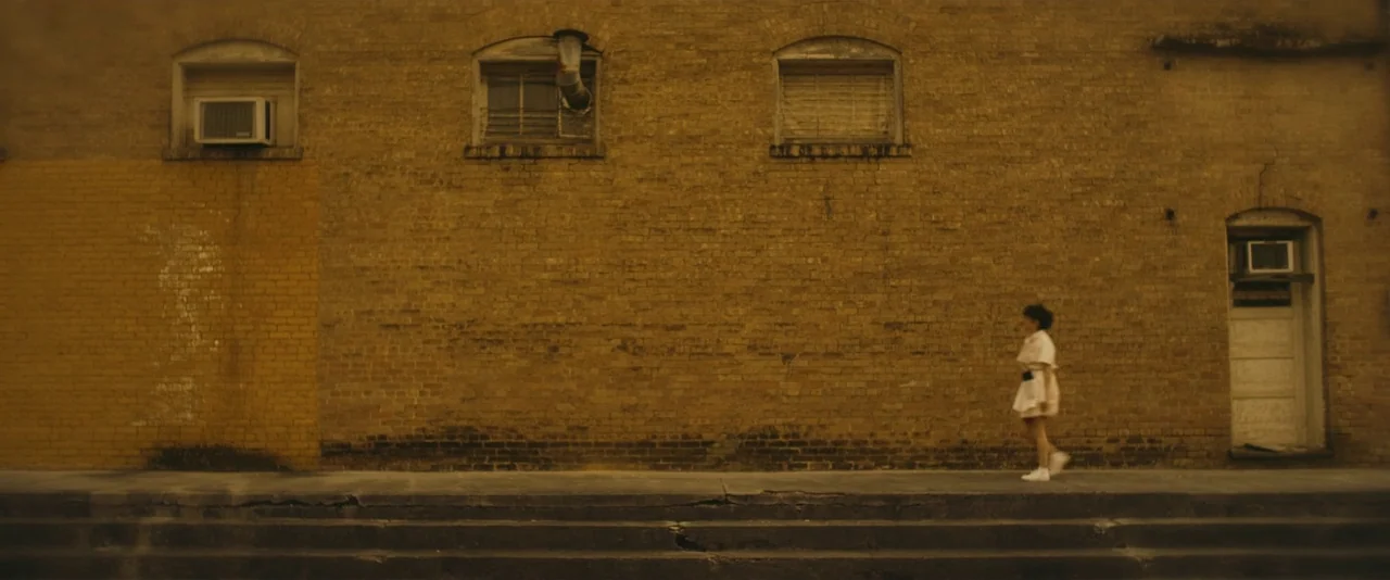 Blaze (2018) movie still: wide — A long yellow-brown brick wall features three windows and an arched doorway entry. A woman walks alone across the…