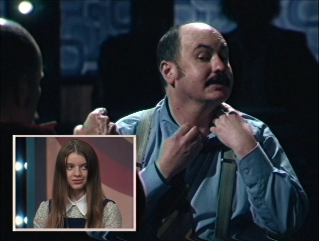 Late Night With The Devil (2023) movie still: medium — A television broadcast set features a man in the foreground and a video inset. The man…