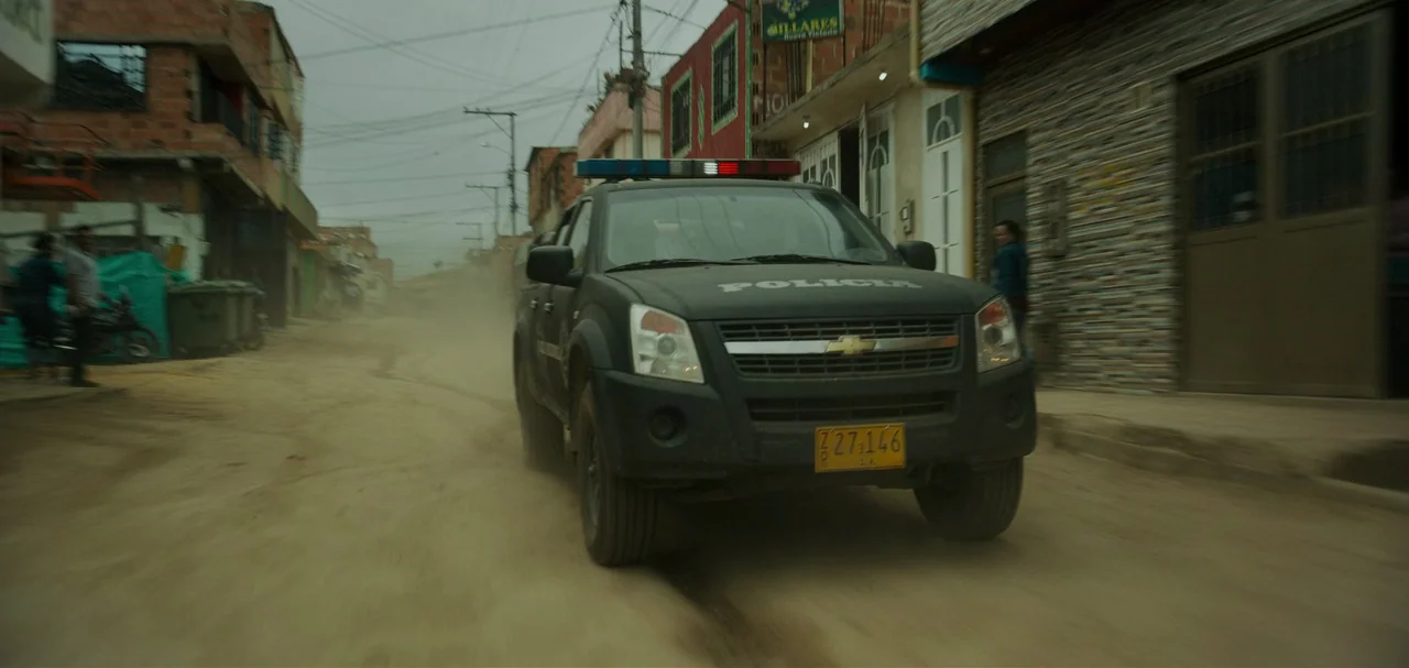 Triple Frontier (2019) movie still: medium — A dark police pickup truck drives down a dusty, narrow road lined with houses. The unpaved street is…