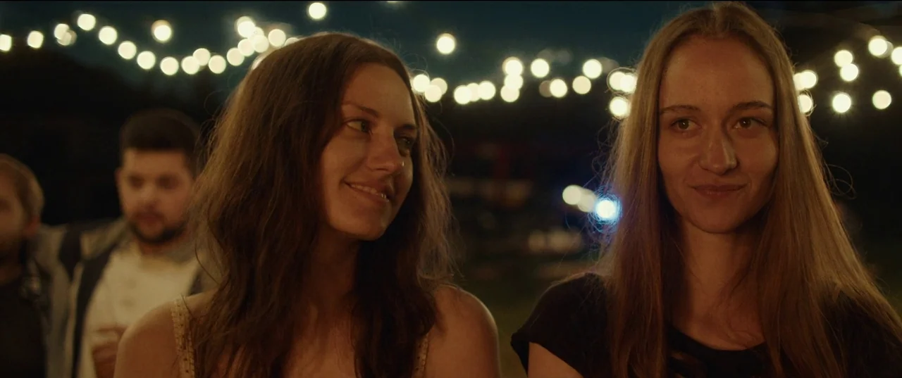 Nightsiren (2022) movie still: medium — An outdoor evening gathering illuminated by glowing strings of hanging festive bulbs. Two women smile at…