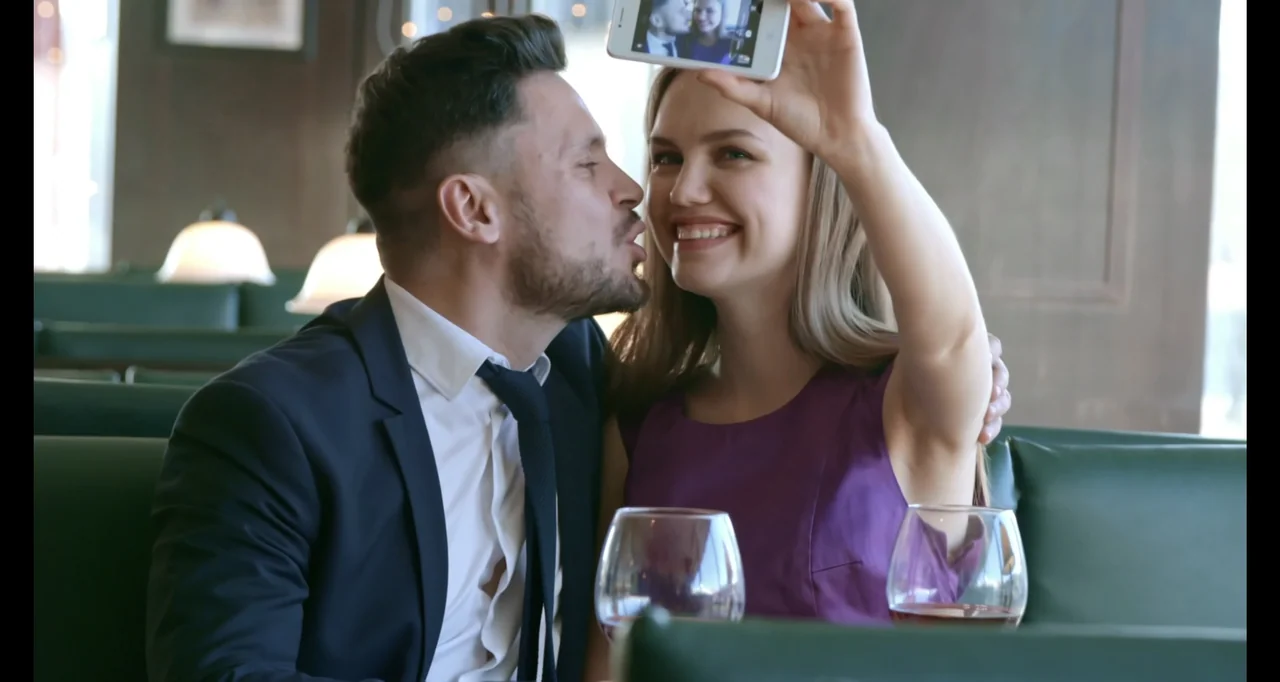 Paired Up (2019) movie still: medium — A man and woman sit together in a green leather booth at a restaurant. The woman holds a white phone up to…