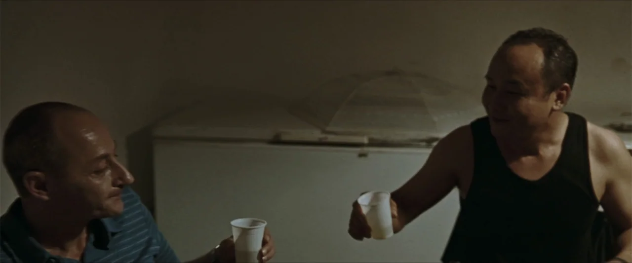 GOMORRAH (2023) movie still: medium — A sparsely furnished room contains a white chest freezer against a plain wall. Two men sit together while…