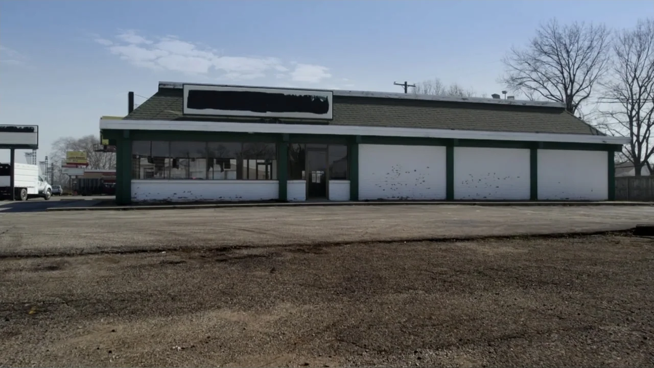 Murders at the Burger Joint (2022) movie still: establishing — A single-story vacant commercial building features white brick walls and dark green…