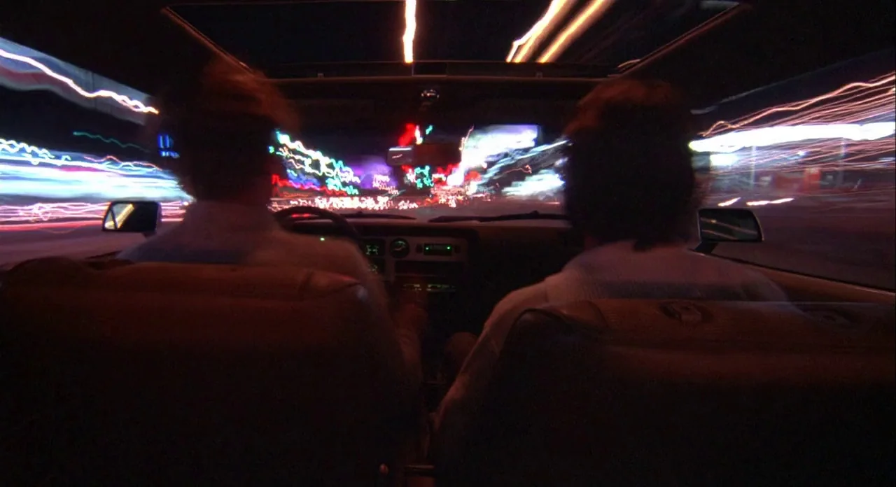 Koyaanasquatsi (1982) movie still: medium — Two people sit in the front seats of a dark moving car at night. Long exposure light trails from city…