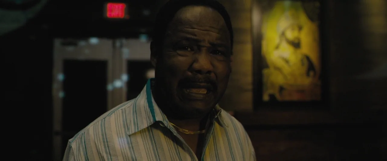 Chi-raq (2015) movie still: medium — A dimly lit room features a framed portrait hanging on a dark wood wall. An older man stands in the foreground…
