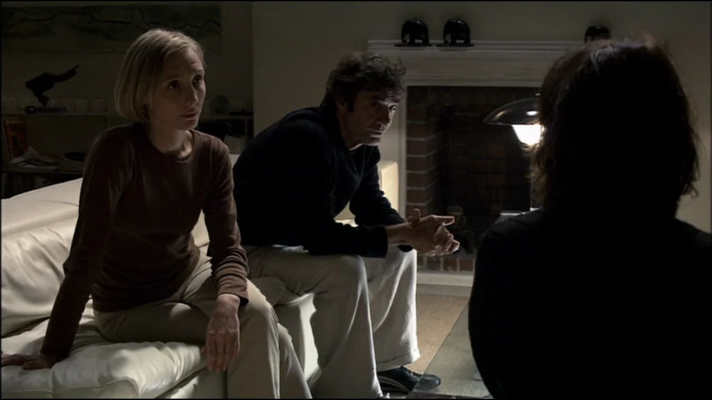 Caché (Hidden) (2005) movie still: medium — A dimly lit room with a fireplace and modern leather seating. A man and a woman sit together on a white…