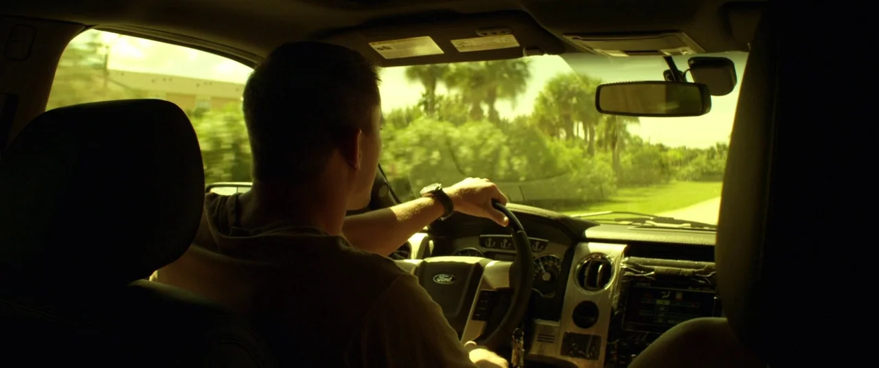 Magic Mike (2012) movie still: medium — A man drives a modern vehicle along a road lined with green tropical trees. The interior of the car is…