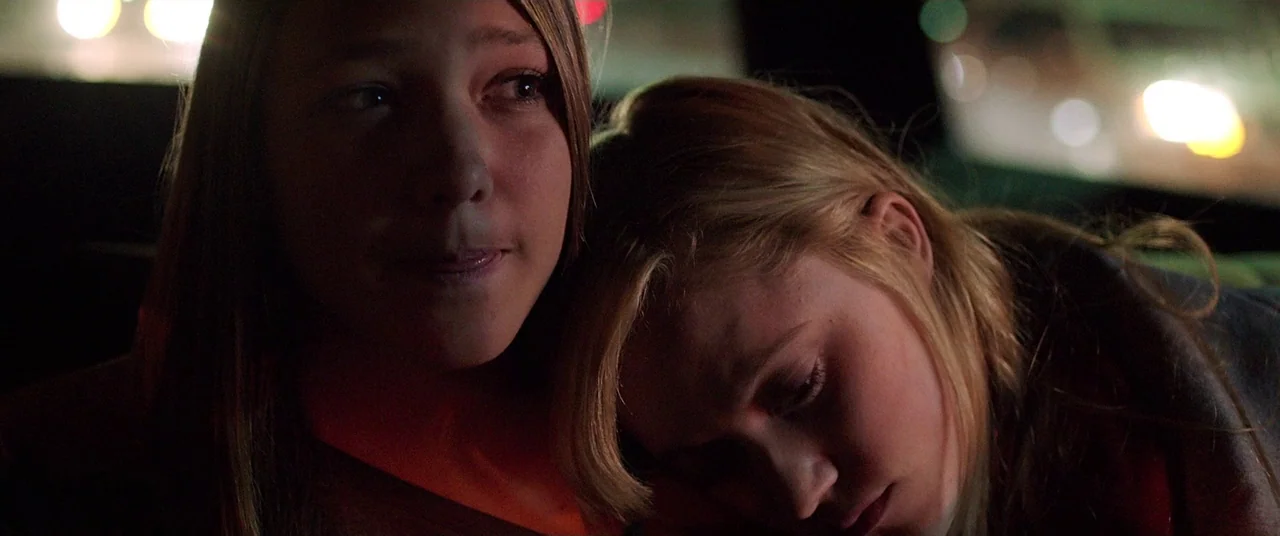 It Follows (2015) movie still: closeup — Inside a dark vehicle, two women are positioned close together in the back seat. One woman stares ahead…