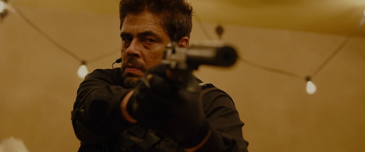 Sicario (2015) movie still: medium — A dimly lit space features suspended string lights against a warm, muted background. A man aims a pistol with a…