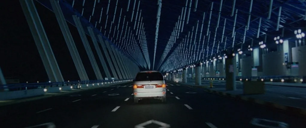 Code 46 movie still: wide — A modern bridge with a distinctive overhead grid structure stretches into the night. A silver minivan drives away from…