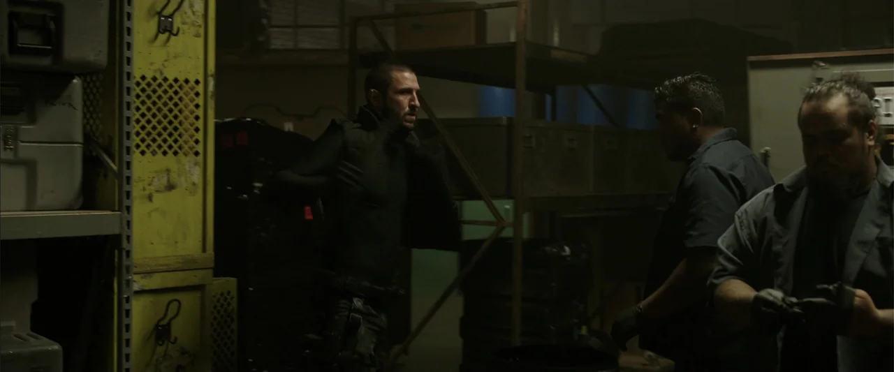 Den of Thieves (2018) movie still: medium — A dimly lit storage area contains yellow metal shelving units and stacked industrial crates. A man in…