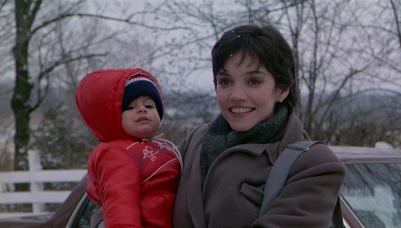 The Dead Zone (1983) movie still: medium — A woman stands outdoors in a winter landscape with bare trees and a white fence. She holds a young child…