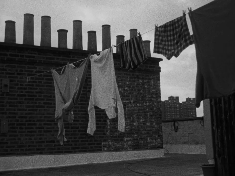 Following (1998) movie still: wide — A rooftop with a red brick wall features several tall cylindrical chimney stacks. Several pieces of clothing…