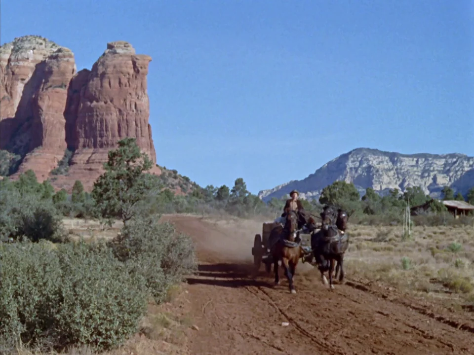 Johnny Guitar (1954) movie still: wide — A dusty dirt track winds through a desert landscape with large red stone formations. A man drives a wagon…